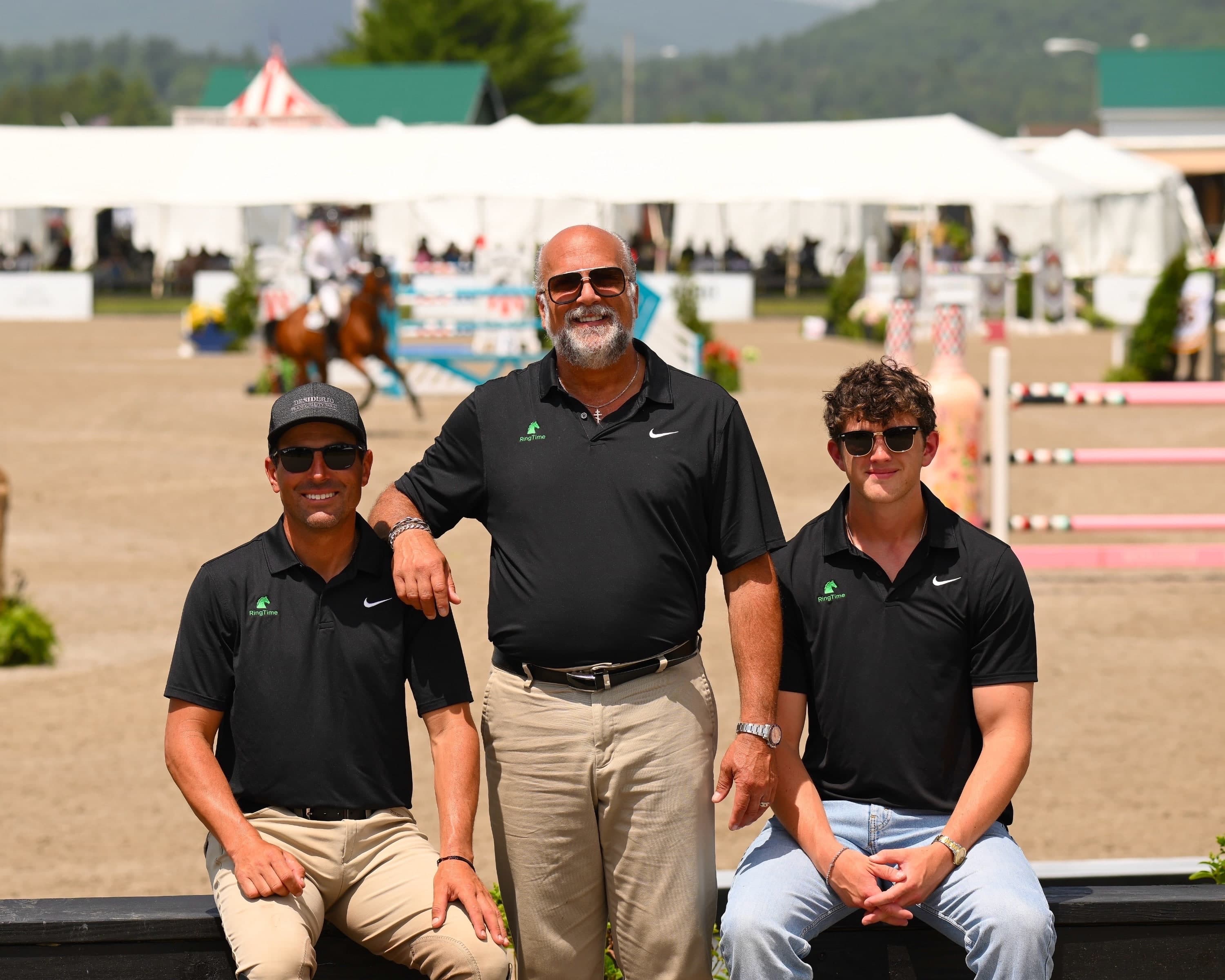 The three RingTime founders in black RingTime polo shirts, two of them sitting on a rail and one standing between them, in front of a sand arena with white tents, jumps and a horse and rider in the background.